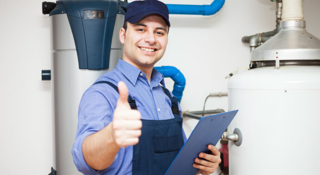 Man in blue cap and plumbing attire giving a thumbs up standing next to a water heater.