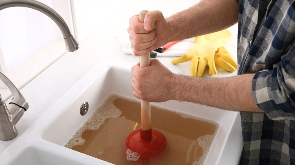 A man using a plunger to unclog a sink against a white countertop and white background.