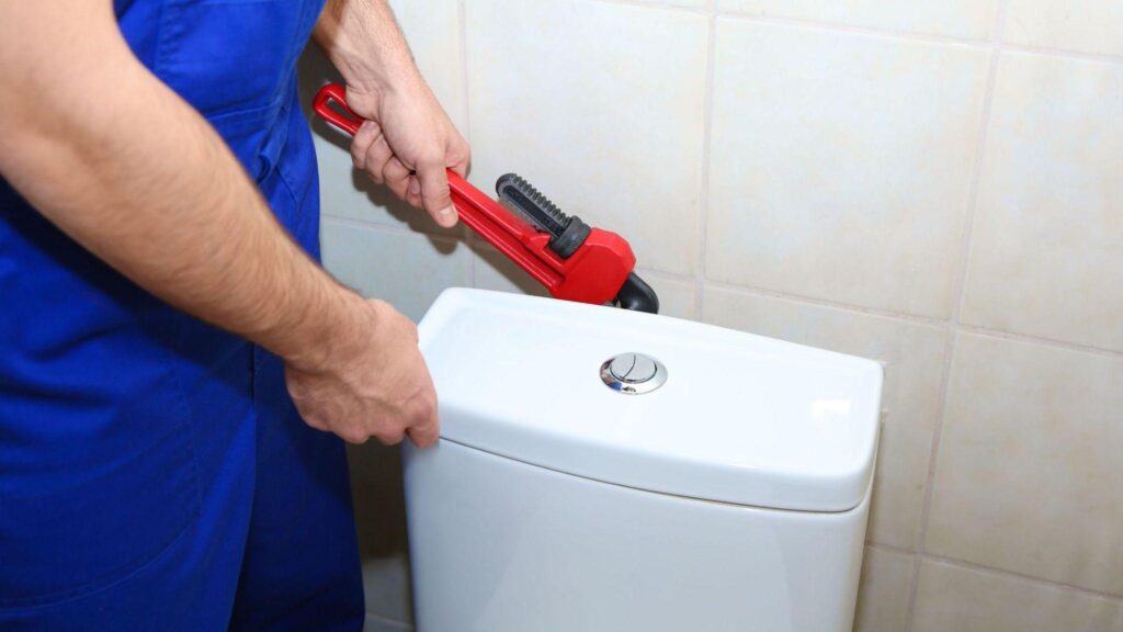 Close up of plumber holding a large red wrench about to open a toilet tank lid.